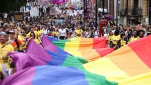 A rainbow pride flag is being held parallel to the floor as marchers in a pride parade move through crowds. 